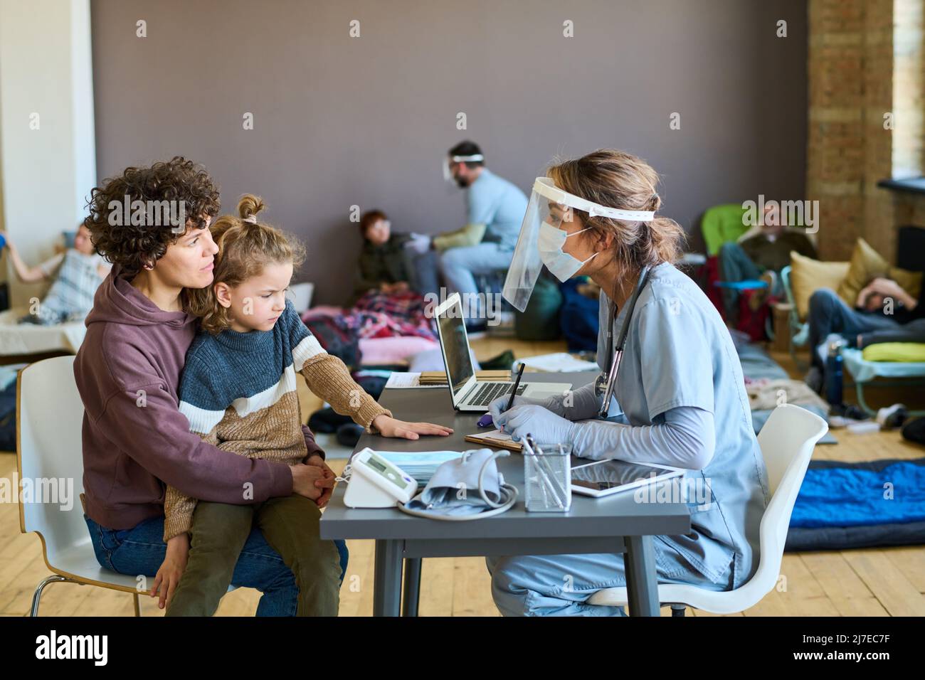 Jeune femme réfugiée avec un petit fils à genoux à l'écoute des conseils médicaux d'un médecin en uniforme, masque de protection et écran Banque D'Images