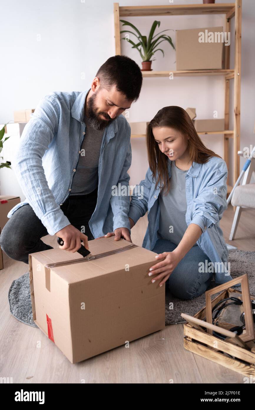 Un homme et une femme le jour du déménagement s'assoient sur le sol et déballent les choses. Un homme coupe le ruban adhésif sur une boîte en carton avec un couteau. Heureux Banque D'Images