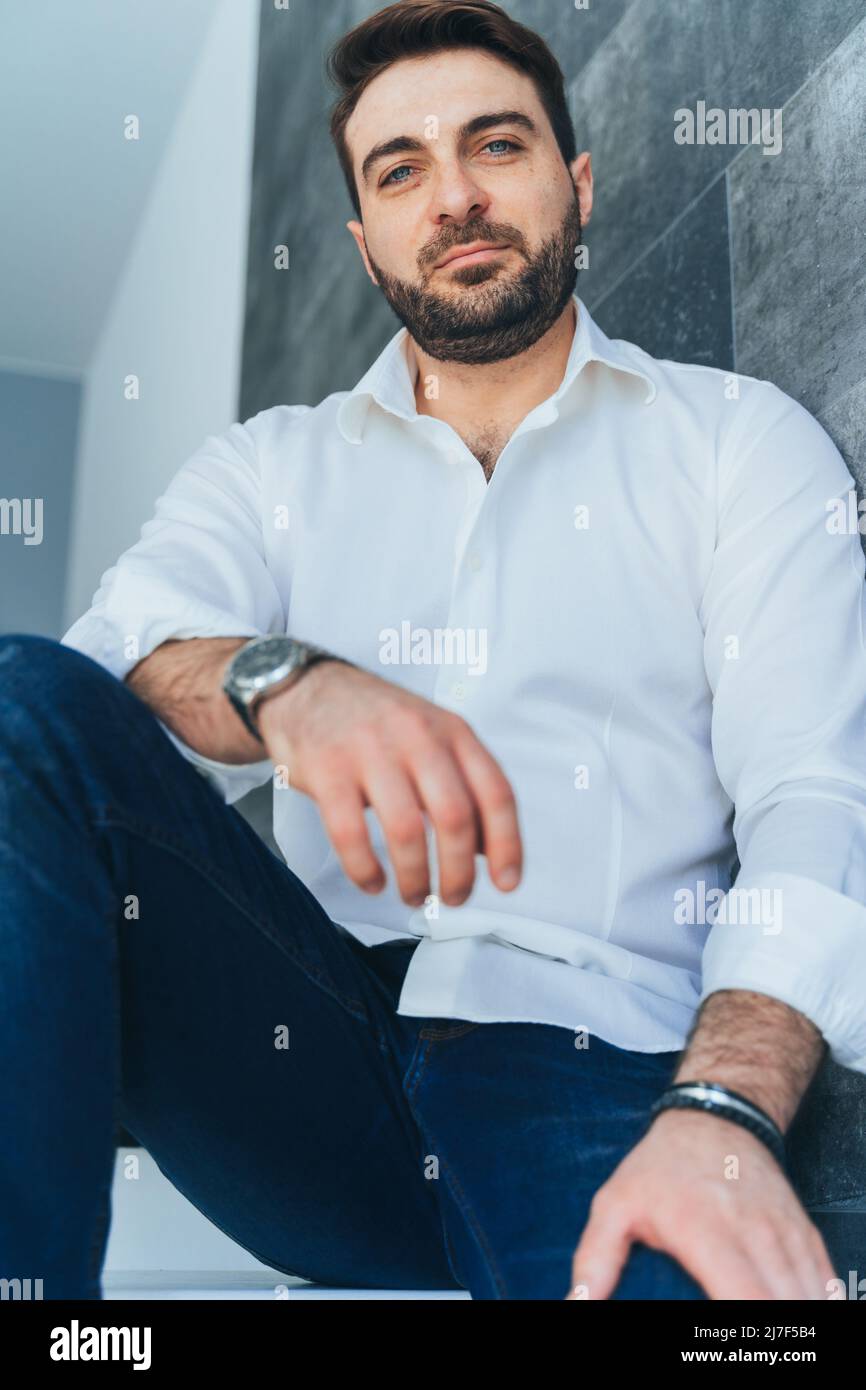 Image verticale de beau et souriant barbu homme, homme d'affaires dans la mode et tendance tenue, regardant l'appareil photo. Assis à l'intérieur, montrant le succès et Banque D'Images