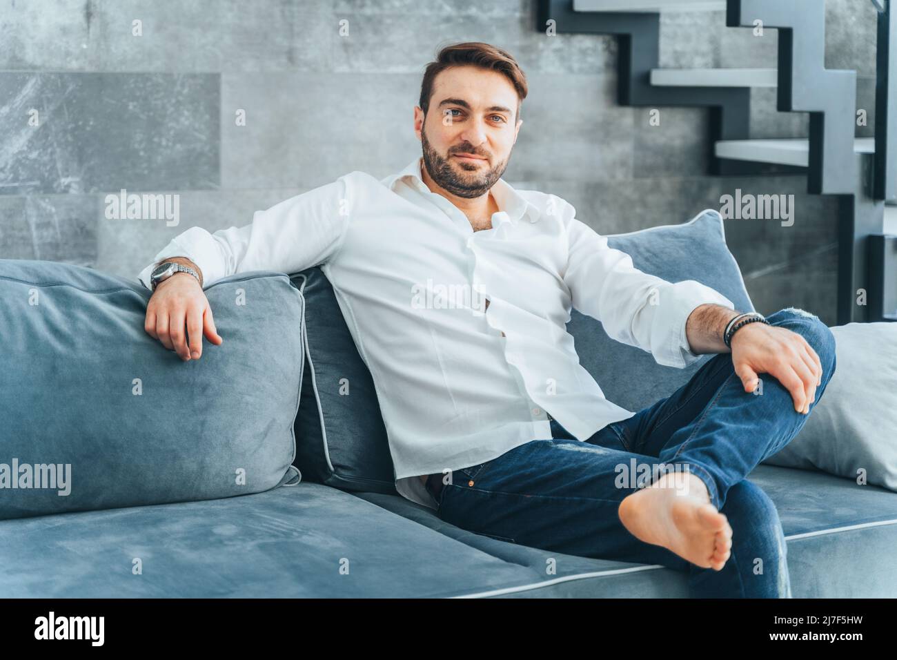 Beau marron, pieds nus, homme d'affaires barbu souriant et confiant regardant la caméra, assis sur un canapé bleu doux à la maison. Repose avec une jambe Banque D'Images