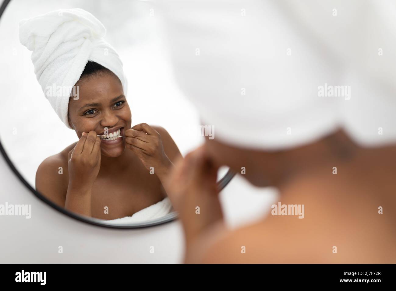 Afro-américaine femme adulte de taille moyenne regardant le miroir en utilisant la soie dentaire dans la salle de bains Banque D'Images