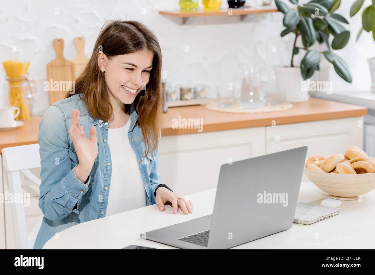 Une femme heureuse et satisfaite travaillant en ligne en utilisant des vagues d'ordinateur portable bonjour à la famille dans la cuisine dans l'appartement moderne de lumière . Style de vie de freelance. À distance Banque D'Images