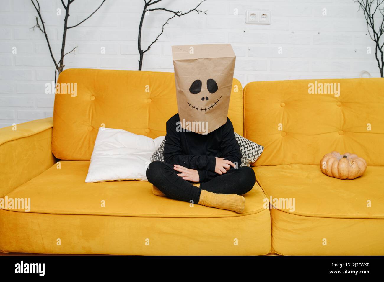 Enfant portant un sac en papier sur sa tête, un visage effrayant est tiré dessus. Assis à pieds croisés sur un canapé orange. Banque D'Images