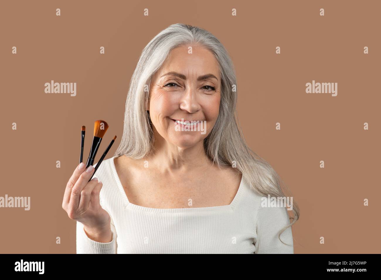 Portrait d'une belle femme mûre tenant un ensemble de différentes brosses cosmétiques sur fond de studio brun Banque D'Images