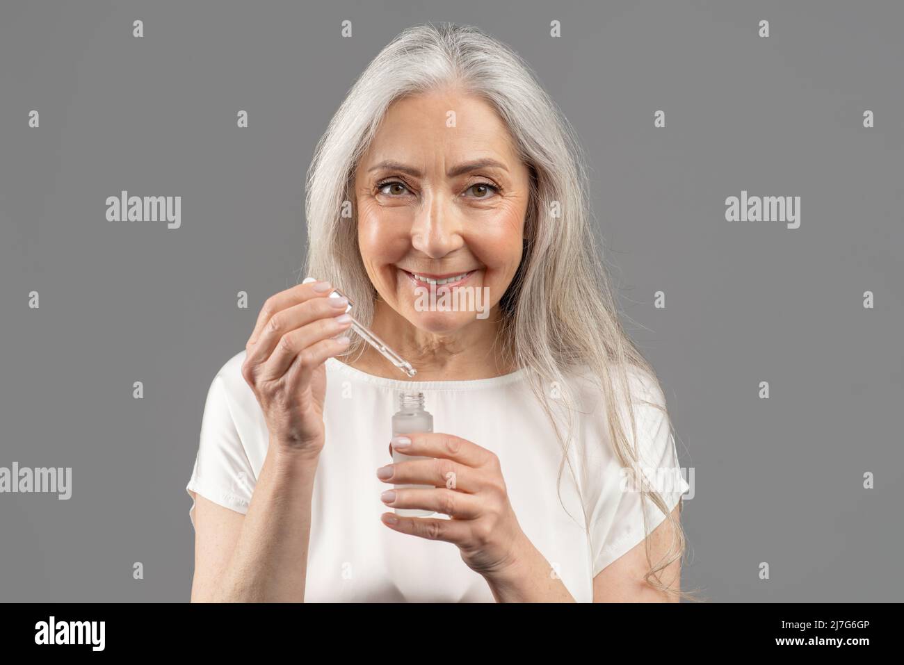 Soins de la peau et bien-être. Portrait d'une femme âgée souriante appliquant du sérum facial avec compte-gouttes sur fond gris de studio Banque D'Images