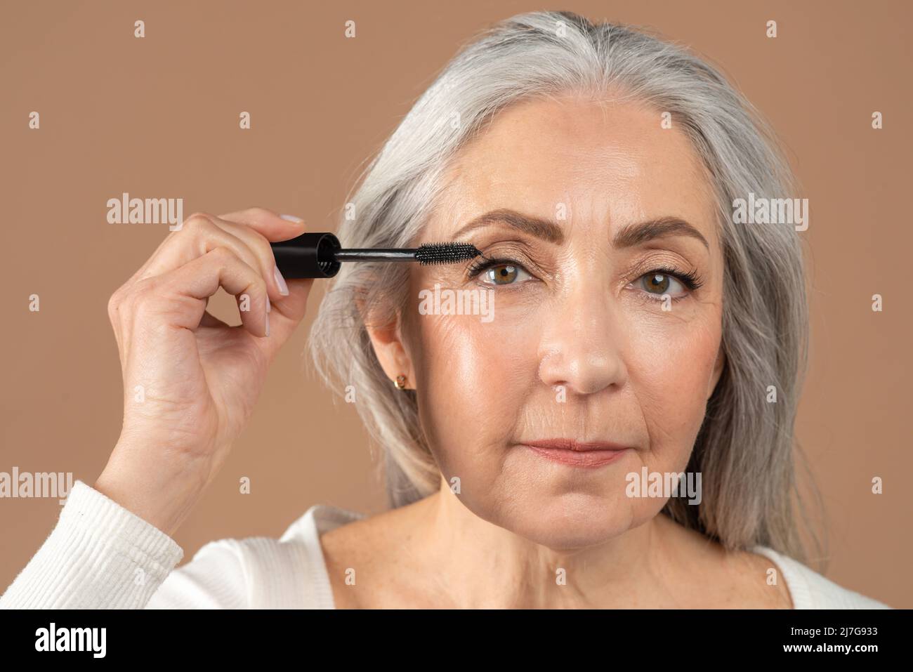 Portrait de beauté de charmante femme âgée appliquant la mascara sur ses cils sur fond de studio brun Banque D'Images