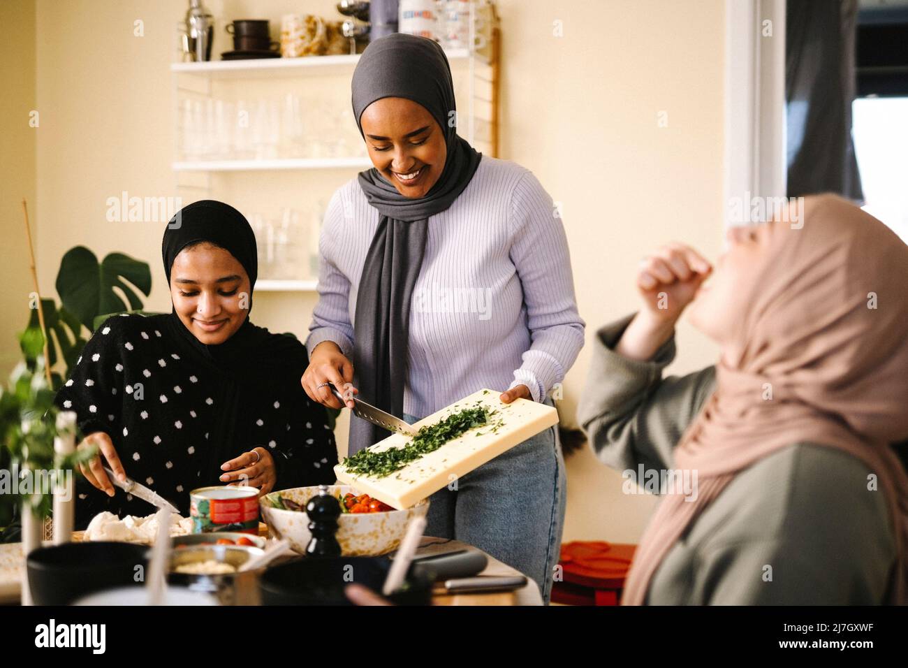 Souriantes, les jeunes femmes hacher des aliments en mangeant par des amis tout en étant assis à une table dans la cuisine Banque D'Images