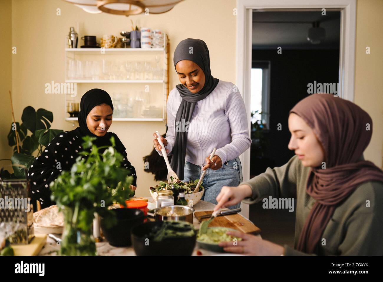 Femme souriante faisant de la salade tout en se tenant debout par des amis préparant la nourriture à table dans la cuisine Banque D'Images