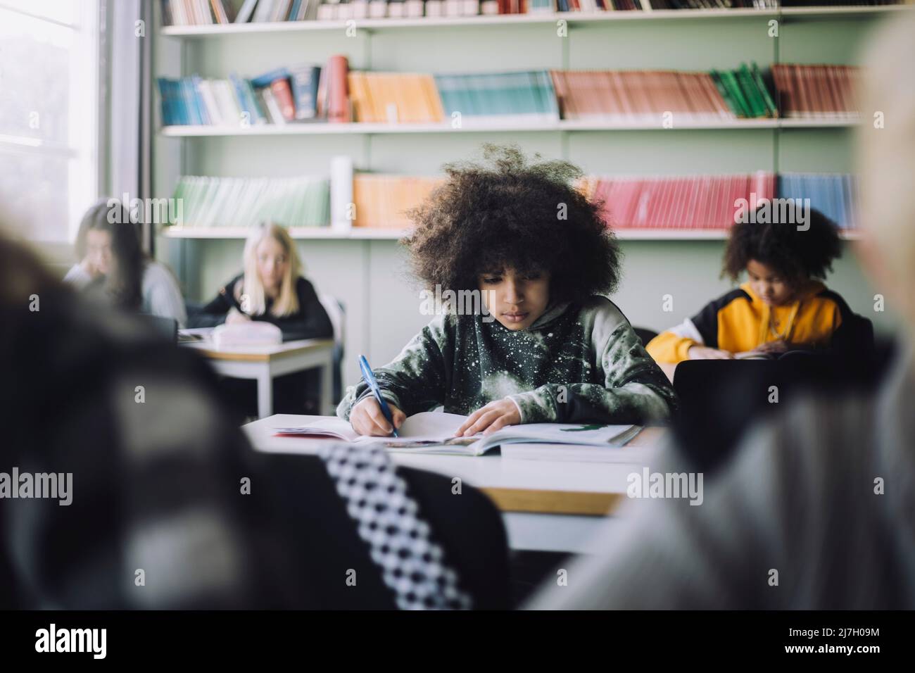 Garçon avec une coiffure afro écrit dans le livre tout en étant assis au bureau dans la salle de classe Banque D'Images