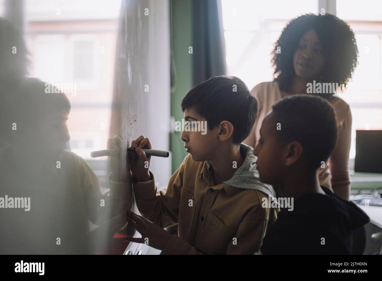 Les garçons font des mathématiques sur le tableau blanc pendant que l'enseignant examine en classe Banque D'Images