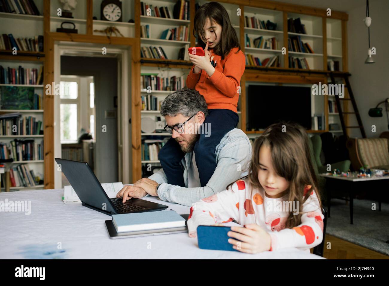 Fille utilisant un smartphone par père travaillant sur ordinateur portable avec une fille assise sur les épaules à la maison Banque D'Images