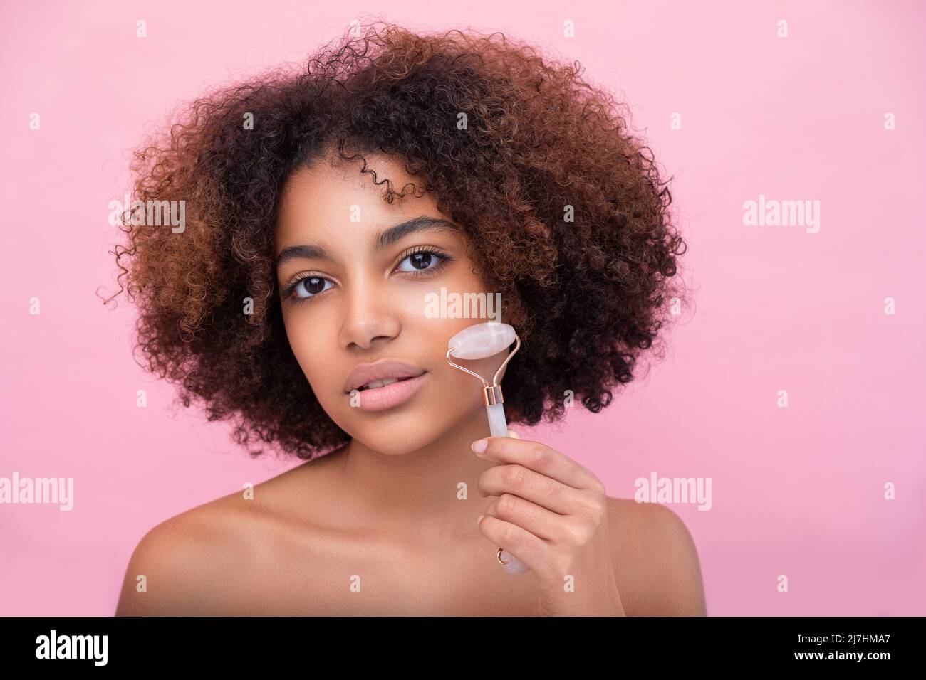 Portrait d'une jeune femme à la peau sombre et bien entretenue avec des cheveux luxuriants, faisant un massage du visage avec l'aide d'un rouleau avec les yeux fermés sur un fond rose gros plan, les soins et les soins de la peau Banque D'Images