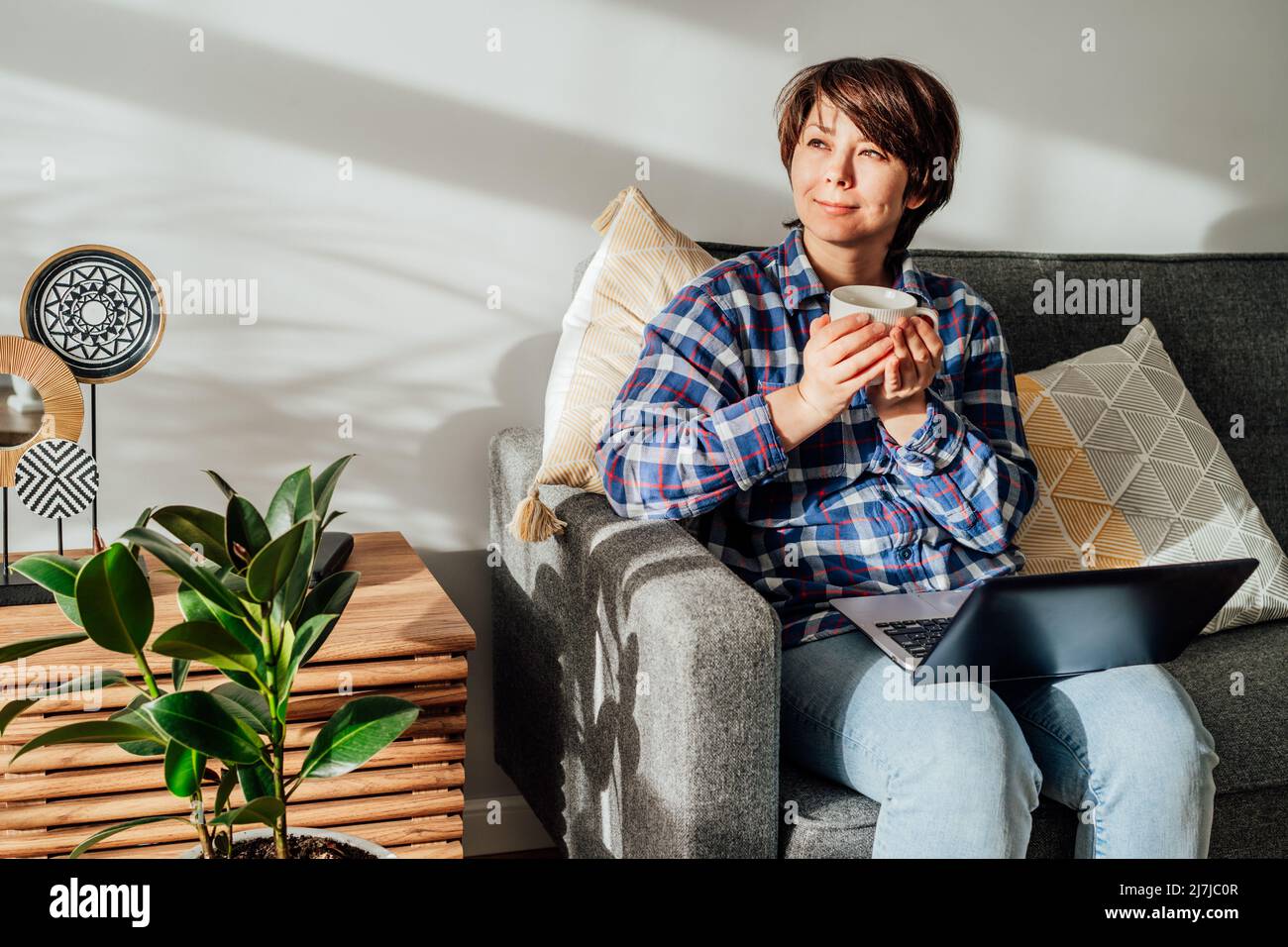Femme rêvant d'utiliser un ordinateur portable et de boire du café, du thé à la maison assis sur un canapé, un canapé aux rayons du soleil. Espace de travail confortable dans un intérieur moderne avec pl. Vert Banque D'Images Femme rêvant d'utiliser un ordinateur portable et de boire du café, du thé à la maison assis sur un canapé, un canapé aux rayons du soleil. Espace de travail confortable dans un intérieur moderne avec pl. Vert Banque D'Images