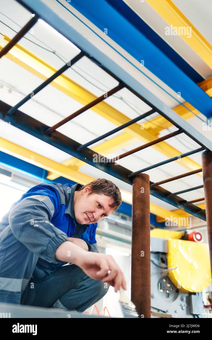 Un jeune travailleur d'apparence caucasienne dans des combinaisons peint soigneusement la structure métallique dans la pièce avec un pinceau. Véritable employé derrière le processus de travail. Main-d'œuvre manuelle. Banque D'Images
