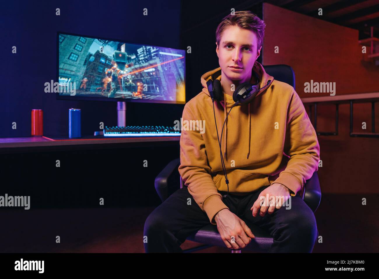 Portrait d'un jeune joueur professionnel sérieux portant un micro-casque assis au bureau avec un équipement de jeu, regardant un appareil photo. Arrière-plan de la pièce de couleur néon. Cybersport, e-sport et jeux vidéo addiction concept Banque D'Images