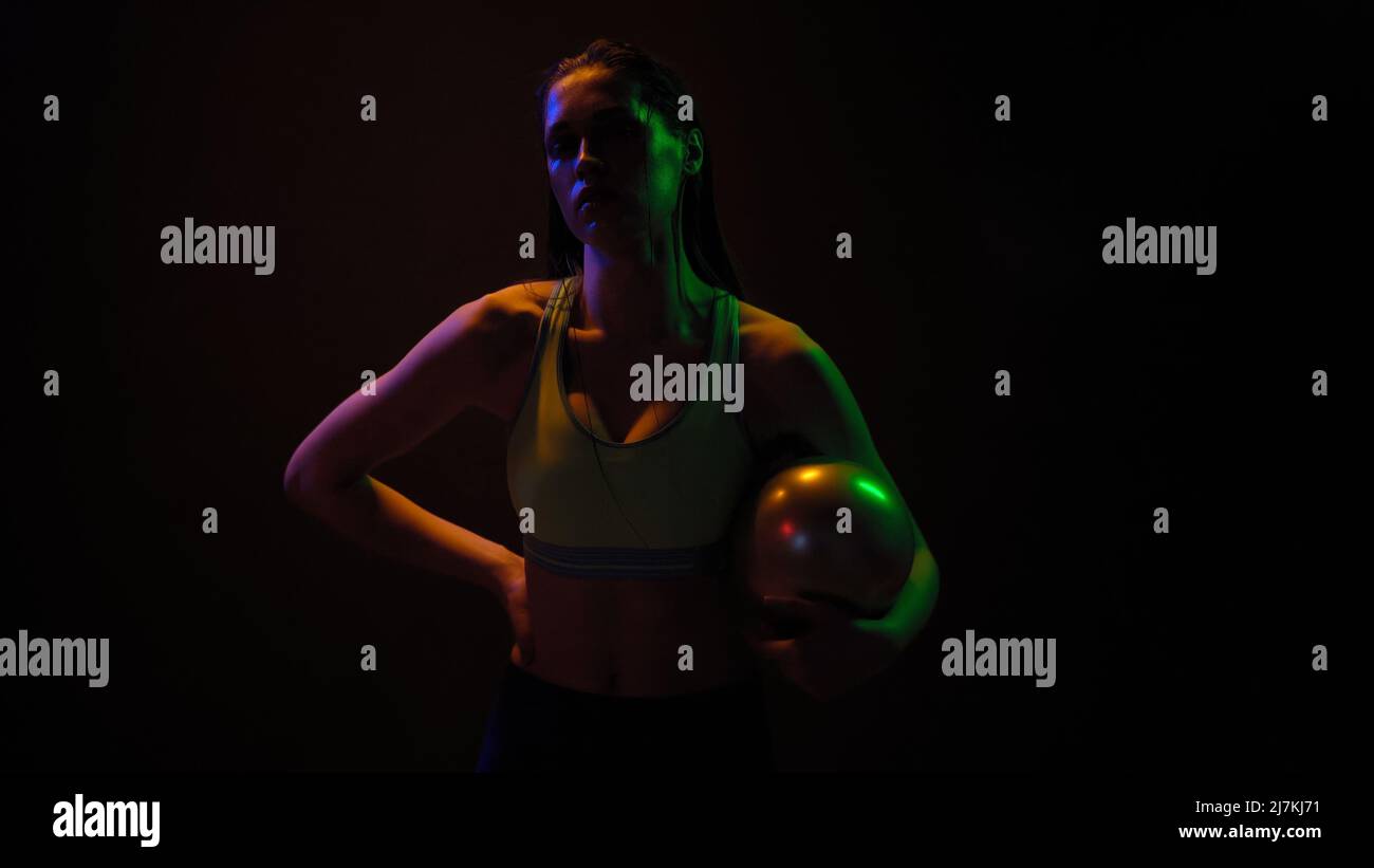 Athlète professionnel fille posant avec la puissance exercice boule debout dans les lumières néon portant sport haut de travail sur un poids fitness isolé sur fond noir. Concept de néon. Banque D'Images