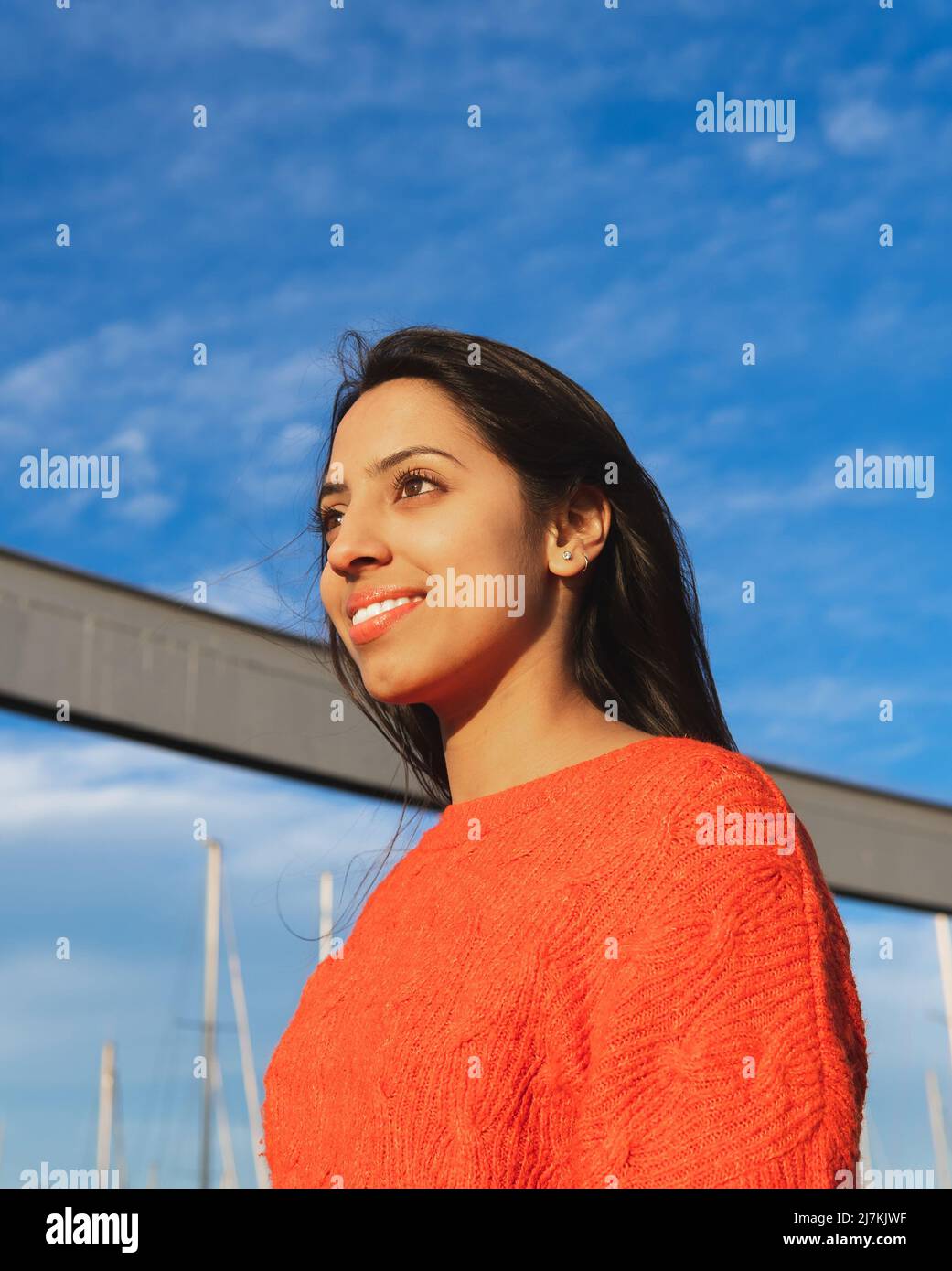 Vue latérale d'une femme indienne positive avec des cheveux noirs en regardant au loin tout en se tenant dans la rue contre le ciel bleu le jour de l'été Banque D'Images