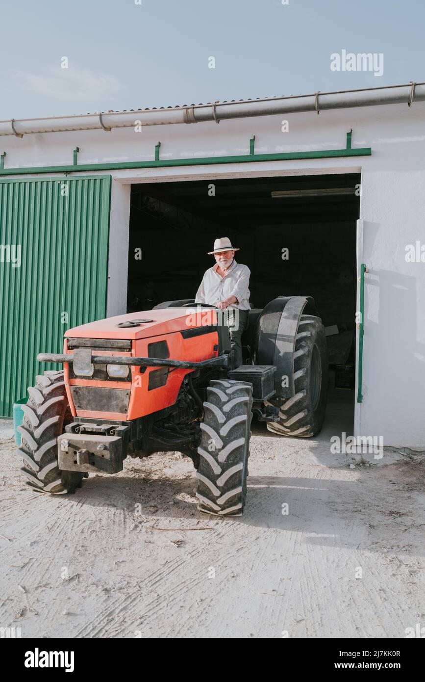 Un fermier en chapeau conduit hors du garage dans un tracteur industriel tout en travaillant dans une zone rurale le jour d'été ensoleillé Banque D'Images