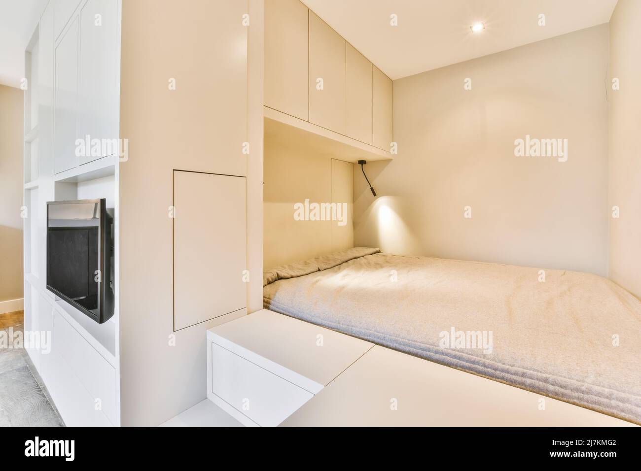 Lit blanc confortable placé au mur près des armoires avec lampe lumineuse placée dans une chambre spacieuse et lumineuse avec télévision moderne dans l'appartement Banque D'Images