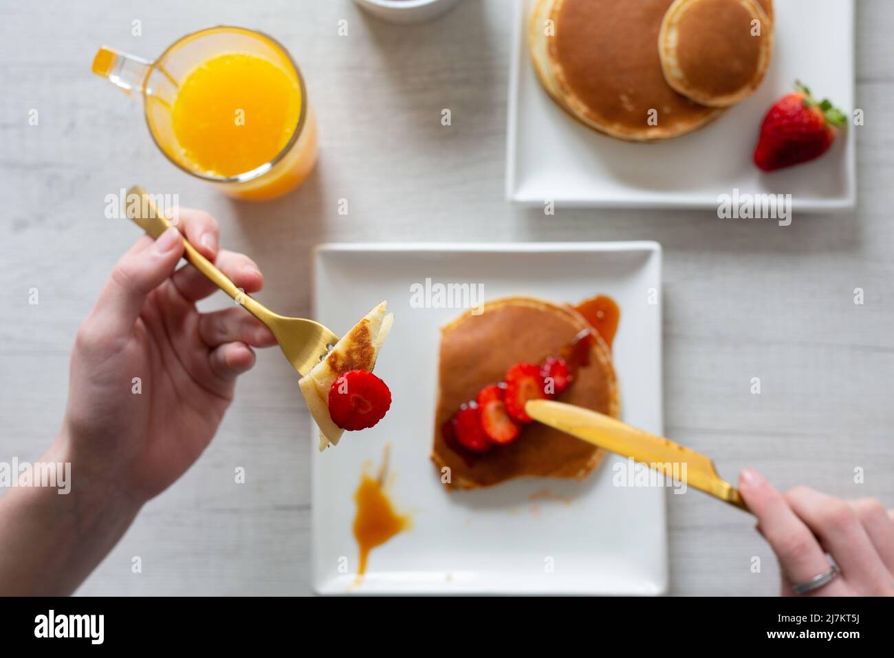 Vue de dessus de crop anonyme personne coupant de savoureuses crêpes sucrées avec des fraises servies sur fond gris avec du jus d'orange dans la cuisine Banque D'Images