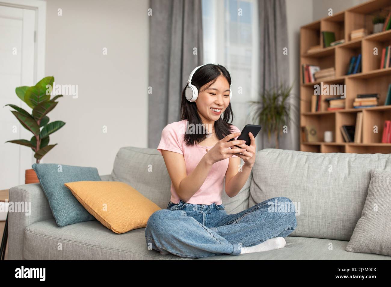 Chat en ligne sur les réseaux sociaux. Bonne jeune femme coréenne dans un casque tapant sur un smartphone, se reposant sur un canapé Banque D'Images