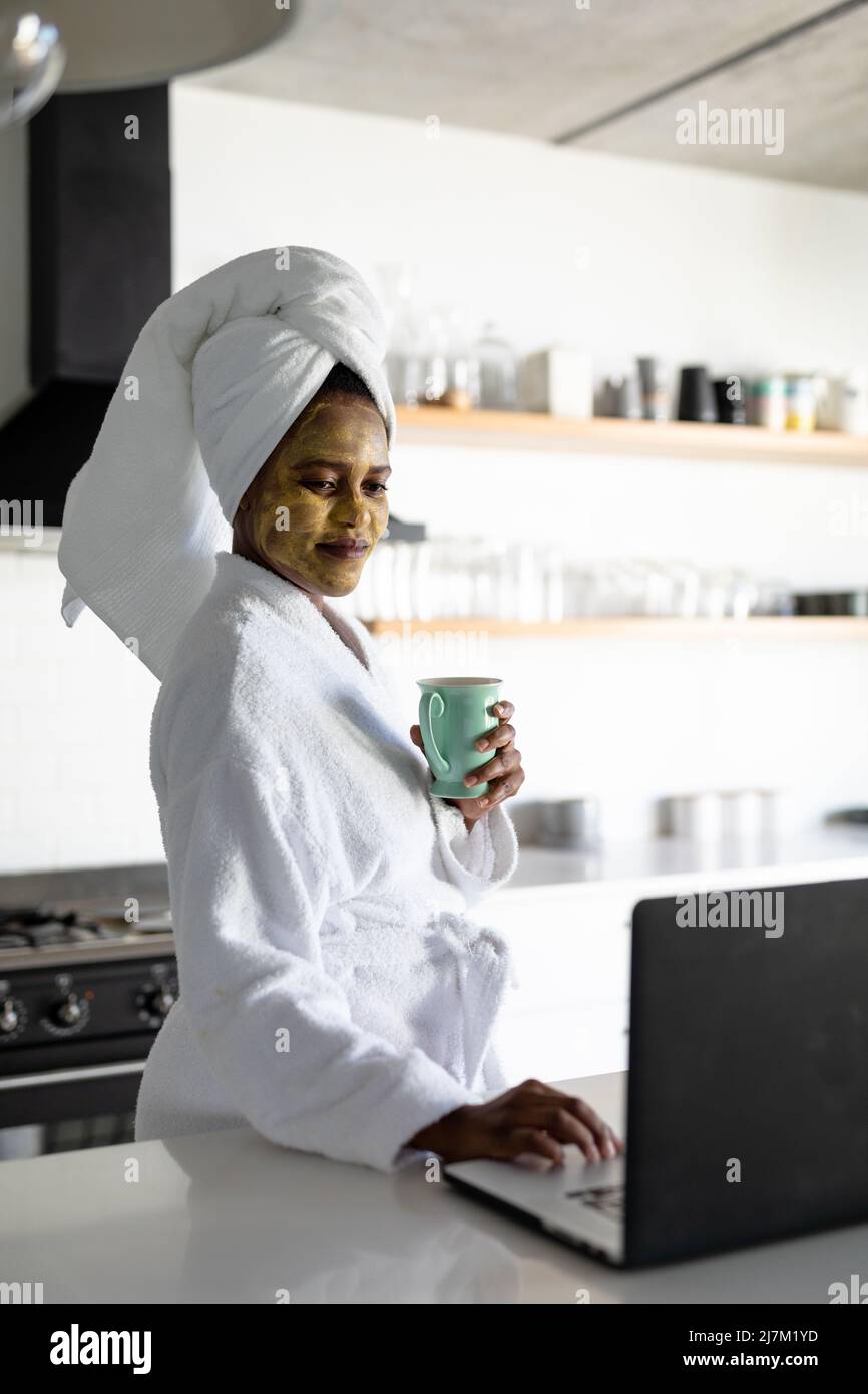 Afro-américaine femme adulte de taille moyenne avec un paquet visage ayant du café et utilisant un ordinateur portable dans la cuisine à la maison Banque D'Images