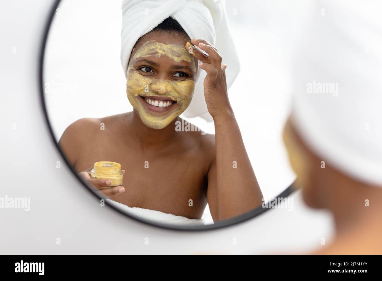 Souriante afro-américaine femme adulte de taille moyenne regardant dans le miroir et appliquant le paquet de visage Banque D'Images