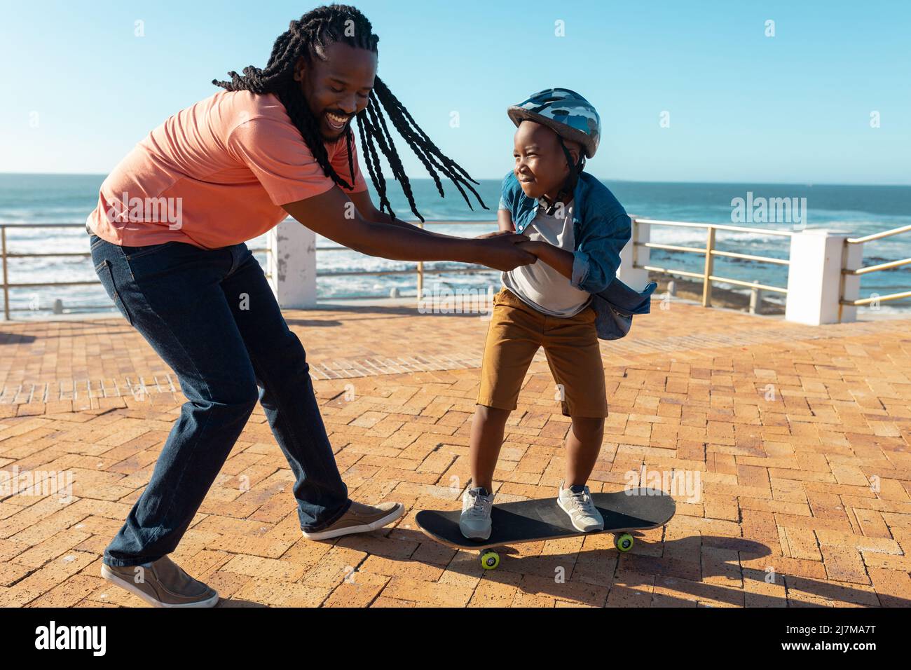 Joyeux père afro-américain aidant son fils à apprendre le skateboard sur la promenade le jour ensoleillé Banque D'Images