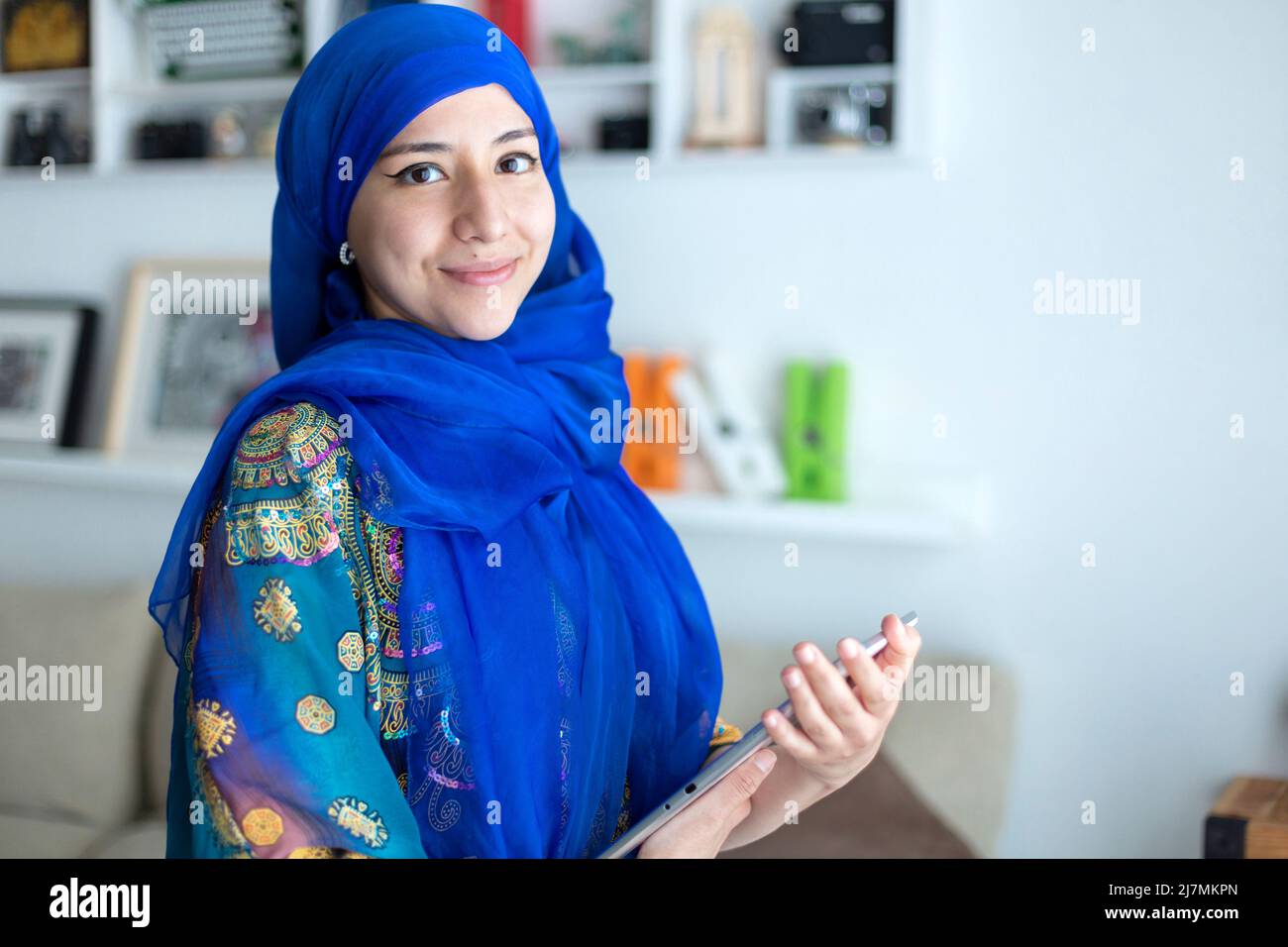 Jeune femme souriante à la maison portant une robe musulmane traditionnelle typique. Elle tient une tablette numérique. Espace pour le texte. Banque D'Images
