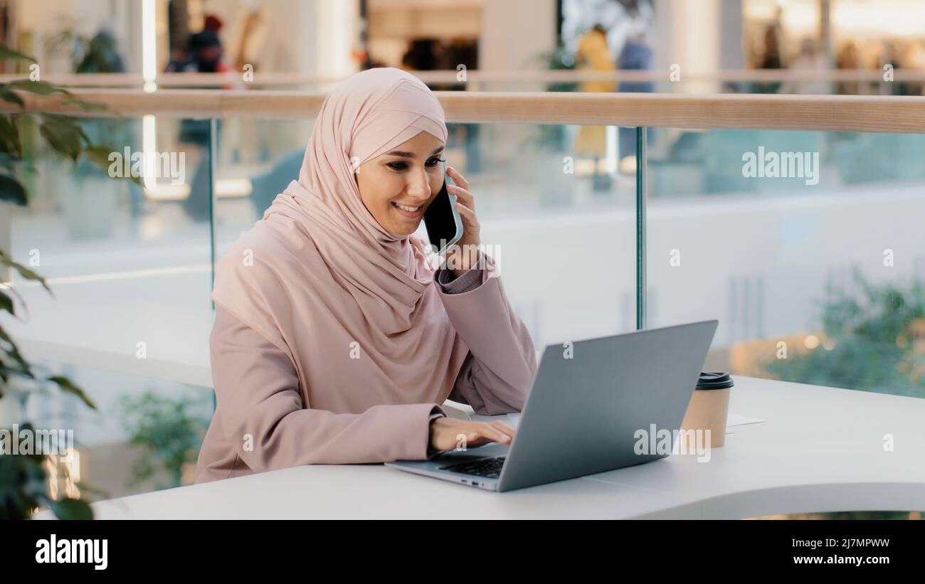 Femme arabe multitâche travaillant sur un ordinateur portable parlant sur un téléphone mobile consultant à distance le client à l'aide d'un smartphone vérifie le réseau social Banque D'Images