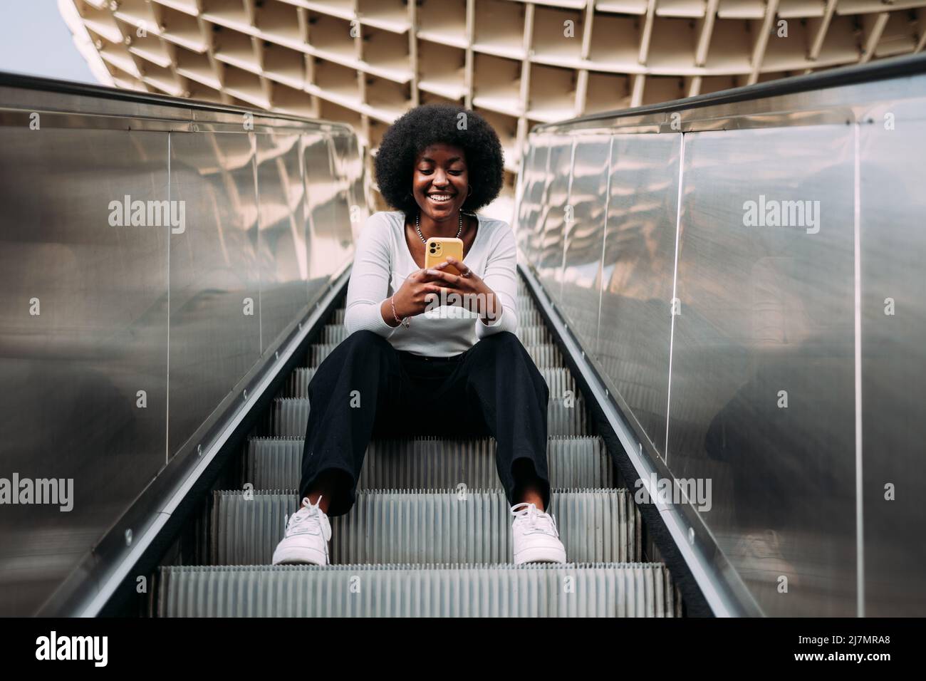 Jeune adolescente noire souriante avec des cheveux afro assis sur un escalator extérieur tapant sur son téléphone. Banque D'Images