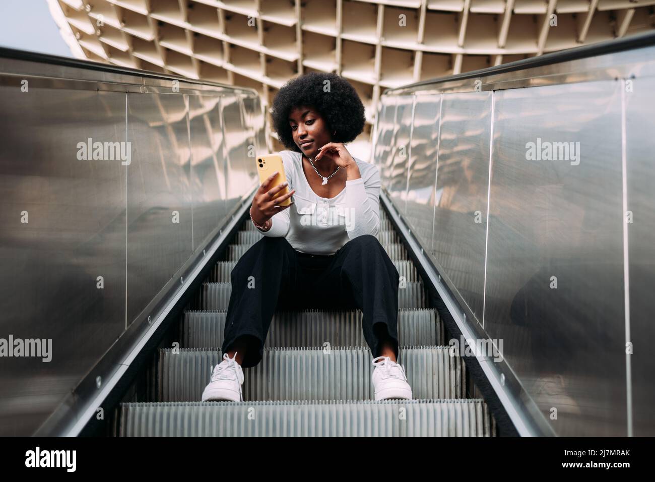 Jeune femme adolescente noire avec cheveux afro assis sur un escalier roulant en plein air prenant un selfie. Banque D'Images