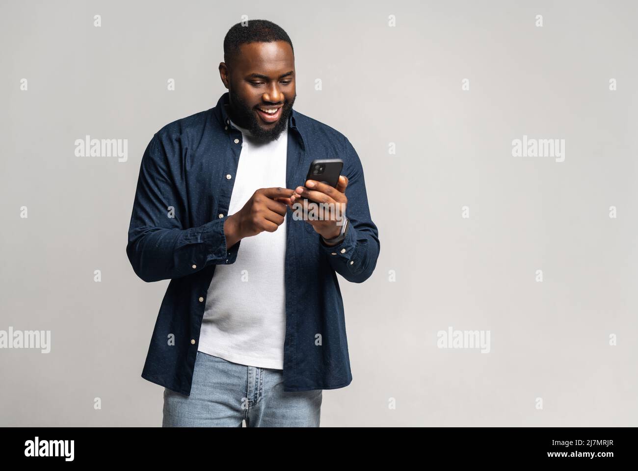 Homme afro-américain heureux dans une chemise décontractée sur un t-shirt avec smartphone isolé sur gris, homme noir chattant en ligne, à l'aide de la nouvelle application mobile, pleine longueur Banque D'Images