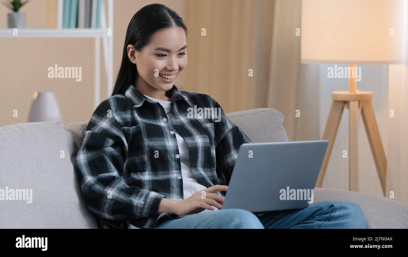 Femme asiatique souriante bonne étudiante fille freelancer assis sur le canapé en utilisant le PC portable dactylographiant le message email dame chat sur l'ordinateur de navigation médias Internet Banque D'Images