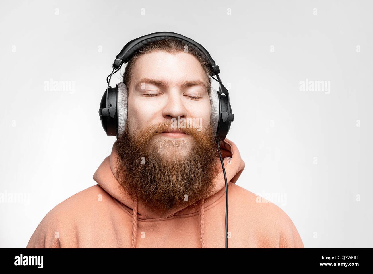 Un homme scandinave charmant et content a fermé les yeux et écoute de la musique dans des écouteurs professionnels isolés sur fond gris. Joyeux gars au gingembre Banque D'Images