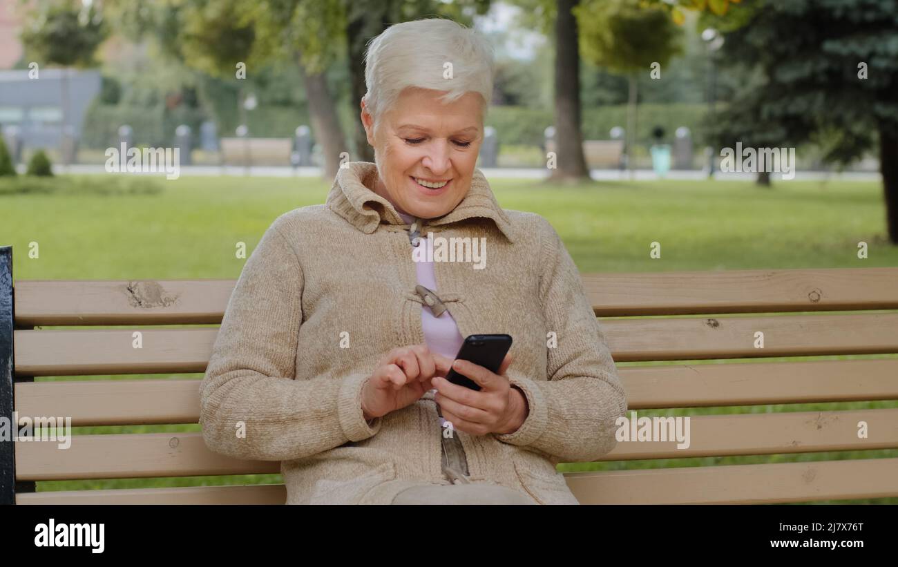 Femme âgée en âge de retraite avec de courts cheveux gris assis dans le parc à l'extérieur tenant le téléphone. Vieille femme élégante souriante regardant des photos sur un smartphone Banque D'Images