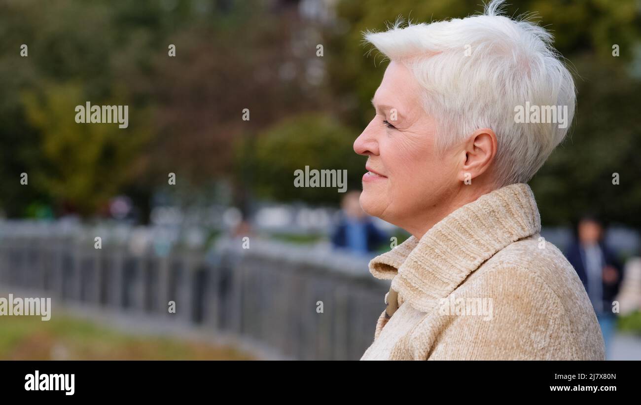 Gros plan portrait 65s bonne femme mûre souriante dans des vêtements décontractés debout posant à l'extérieur tourne la tête regardant de côté. Calme attirante femme bienveillante Banque D'Images