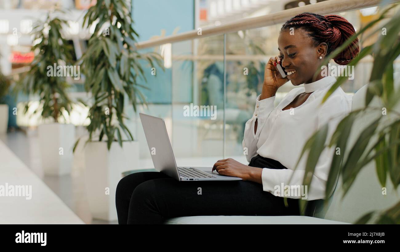 Une jeune fille africaine étudiante assise sur un canapé dans un bureau dans un centre commercial à l'intérieur parlant sur un téléphone mobile répondre à un appel dactylographiant sur un ordinateur portable travail à distance Banque D'Images