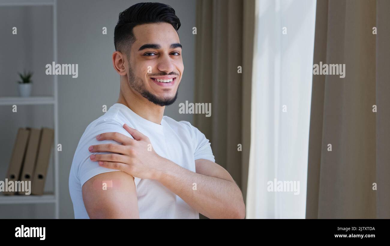 Heureux satisfait indien gars arabe patient hispanique homme posant à l'intérieur porte t-shirt blanc montre le plâtre sur l'épaule se sent soulagement après la vaccination Banque D'Images