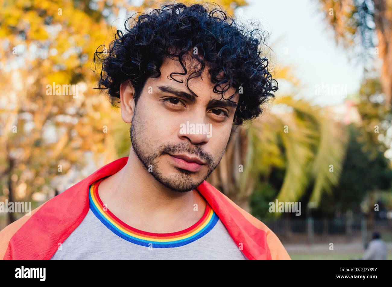 portrait homme caucasien, avec la barbe et les cheveux bouclés, à l'extérieur par temps ensoleillé, avec un drapeau de fierté gay sur ses épaules, regardant l'appareil photo Banque D'Images