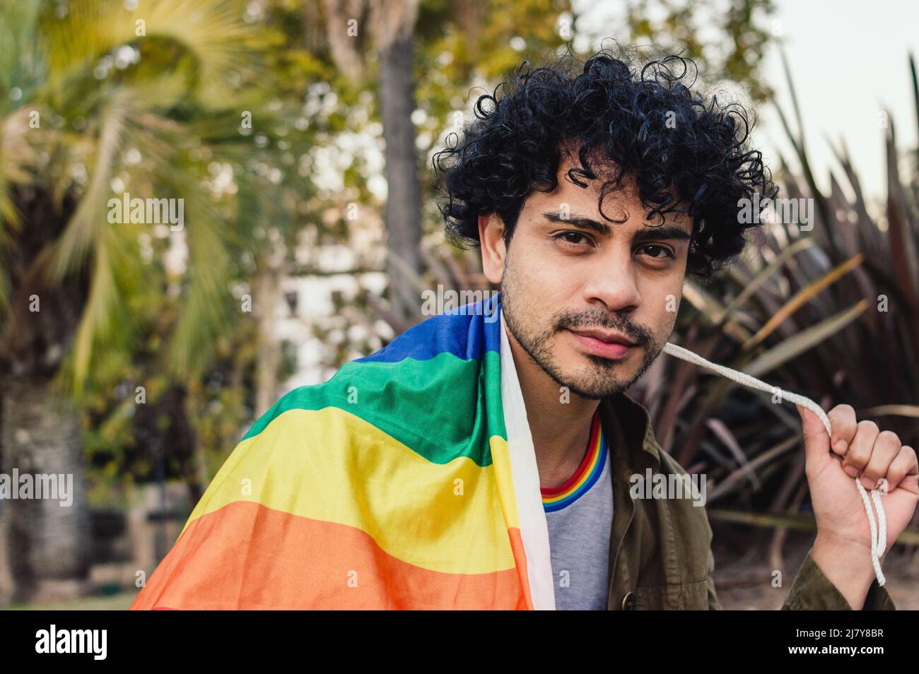 jeune homme caucasien, avec des boucles et une barbe, plein air souriant enveloppé de fierté drapeau regardant l'appareil photo, espace de copie. Banque D'Images