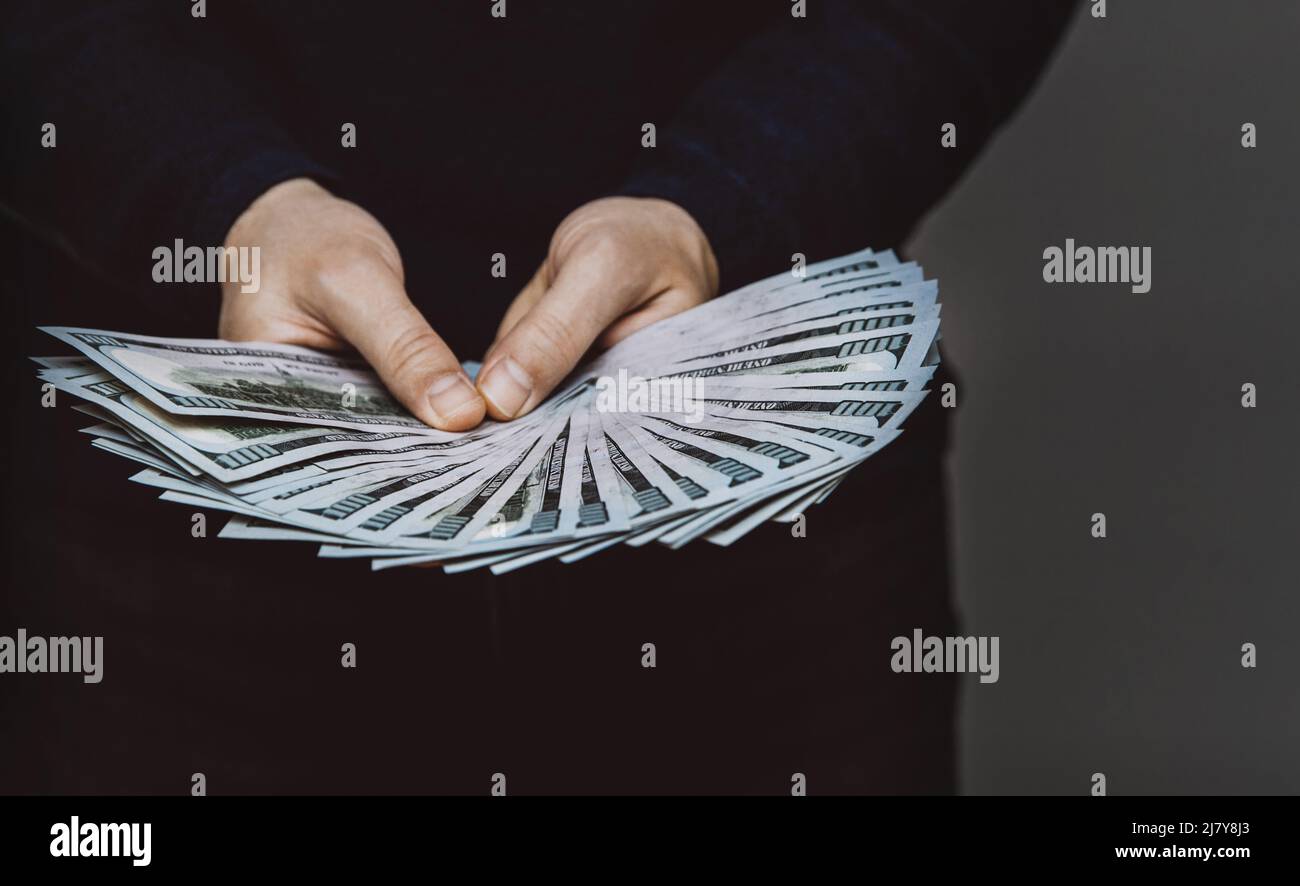 Un homme tient un fan de 100 billets de dollars dans ses mains dans des couleurs sombres. Le concept de revenus durs et de travail sale Banque D'Images
