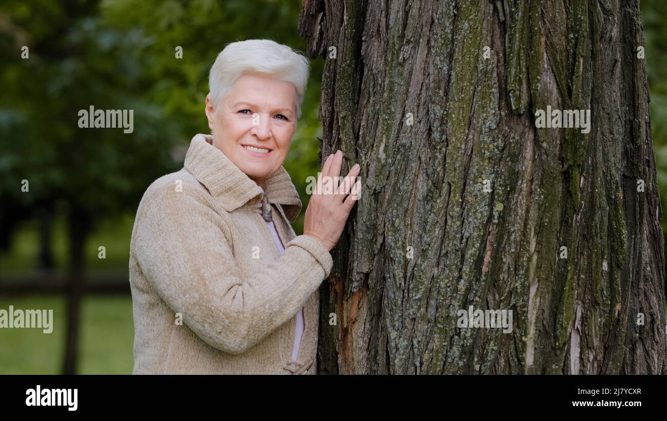 Femme élégante en âge de retraite, se reposant dans le parc seul, se tenant près d'un grand arbre, le caressant, touche tendinement l'écorce. Attitude respectueuse et amour nature Banque D'Images