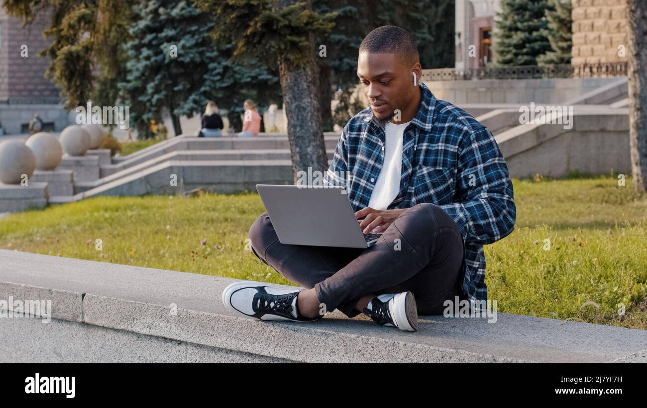 Un étudiant américain, un homme d'affaires africain, assis à l'extérieur dans une rue urbaine en train de taper sur un ordinateur portable, travaillant en ligne avec l'e-learning sur ordinateur à l'écoute de la musique Banque D'Images