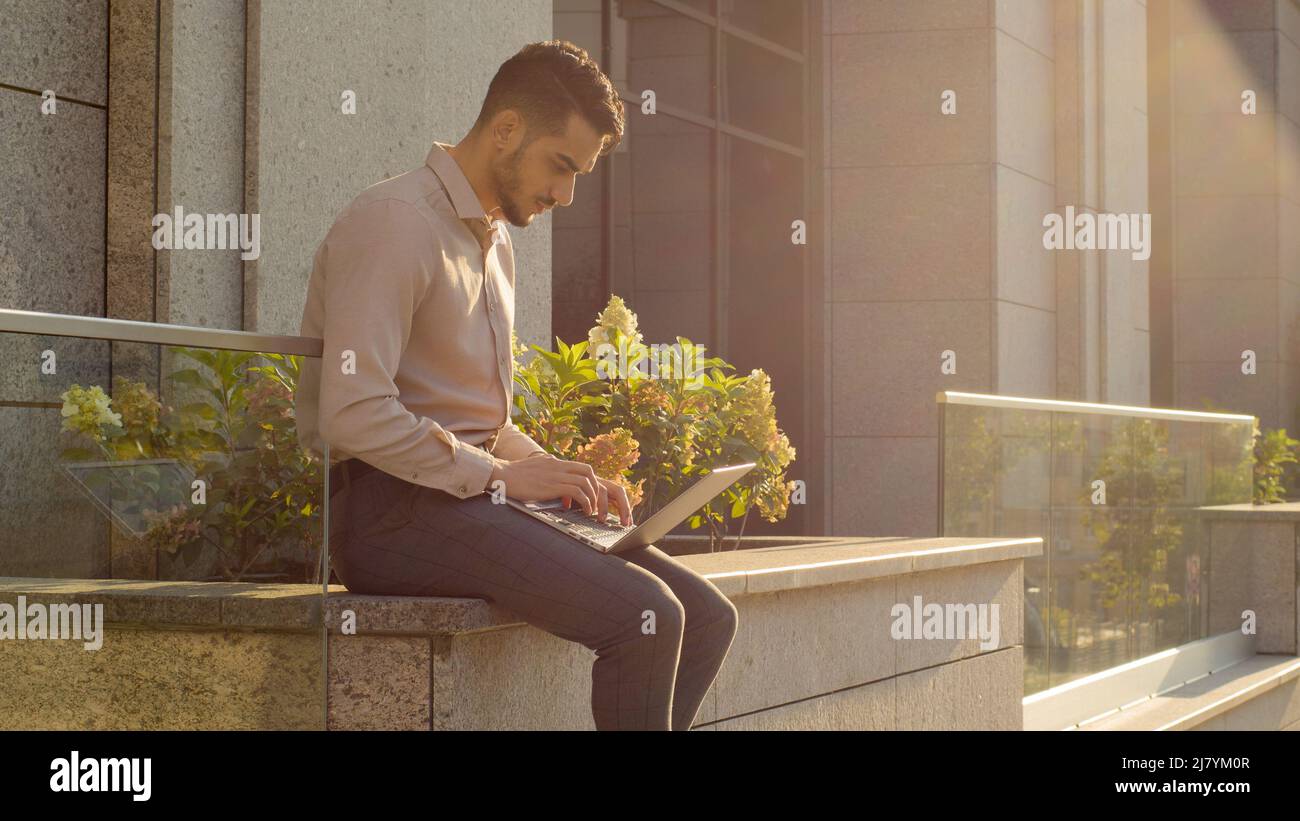 Homme d'affaires hispanique assis à l'extérieur avec un ordinateur portable travaillant à distance travail à l'extérieur près du bâtiment de l'entreprise de bureau. Jeune travailleur étudiant utilisateur arabe Banque D'Images