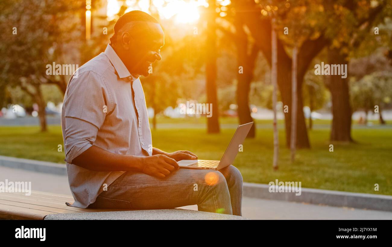 Homme afro-américain âgé assis à l'extérieur, parcourant Internet sans fil sur un appareil moderne, tapant des SMS sur un ordinateur portable travaillant sur un ordinateur, adulte Banque D'Images