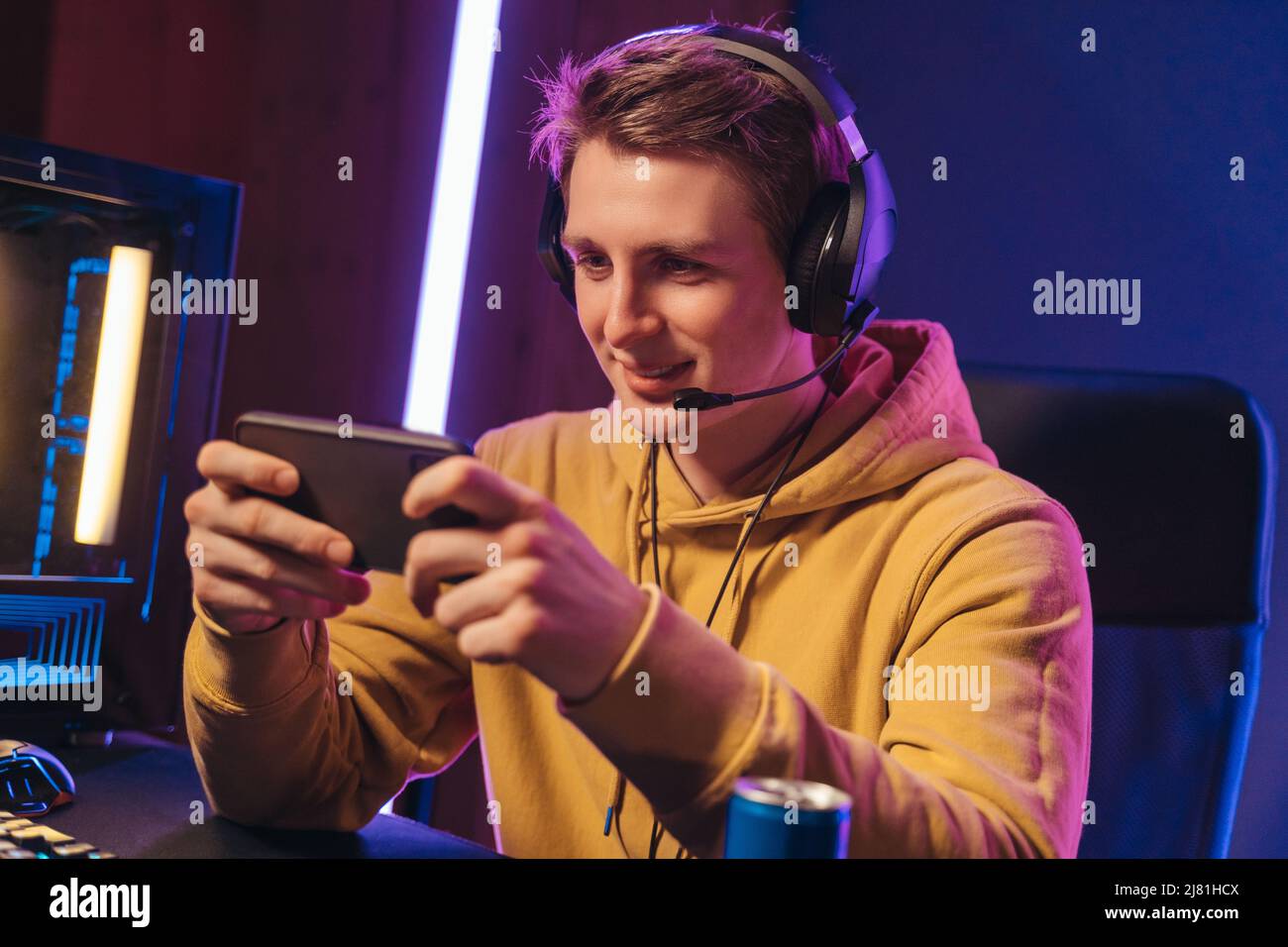 Jeune gamer professionnel de cybersport jouant à un jeu mobile par smartphone sur un tournoi en ligne dans la salle Neon Lights. Un joueur professionnel souriant portant un casque en streaming lorsqu'il participe à un événement eSport Banque D'Images