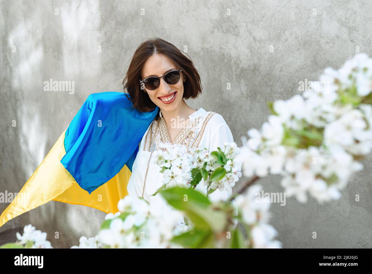 Une fille en robe blanche et des lunettes de soleil en ville sur fond de  mur de béton agite le drapeau ukrainien national, célébrant la victoire.  Fleurs de pomme. Copier l'espace. Idée