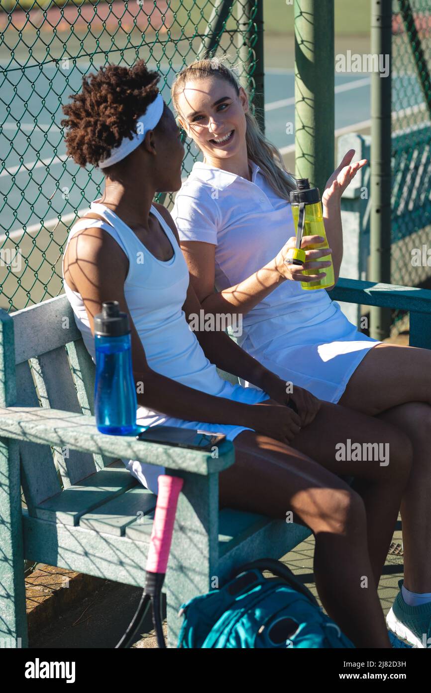 Souriant, les jeunes joueuses de tennis multiraciales parlent tout en étant assise sur le court pendant la pause Banque D'Images