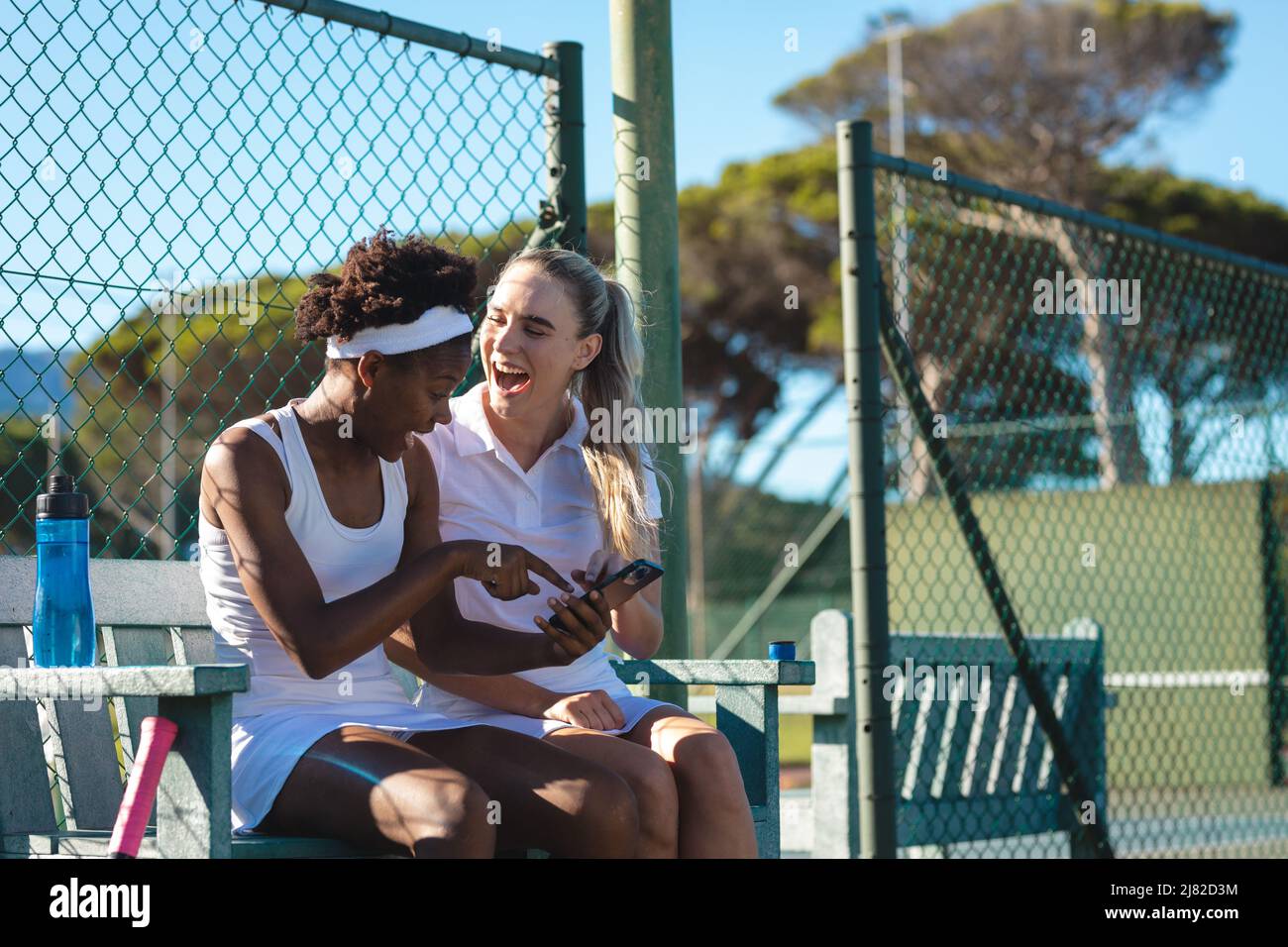Joueuses de tennis biraciales gaies partageant un smartphone tout en étant assis sur le banc au court Banque D'Images